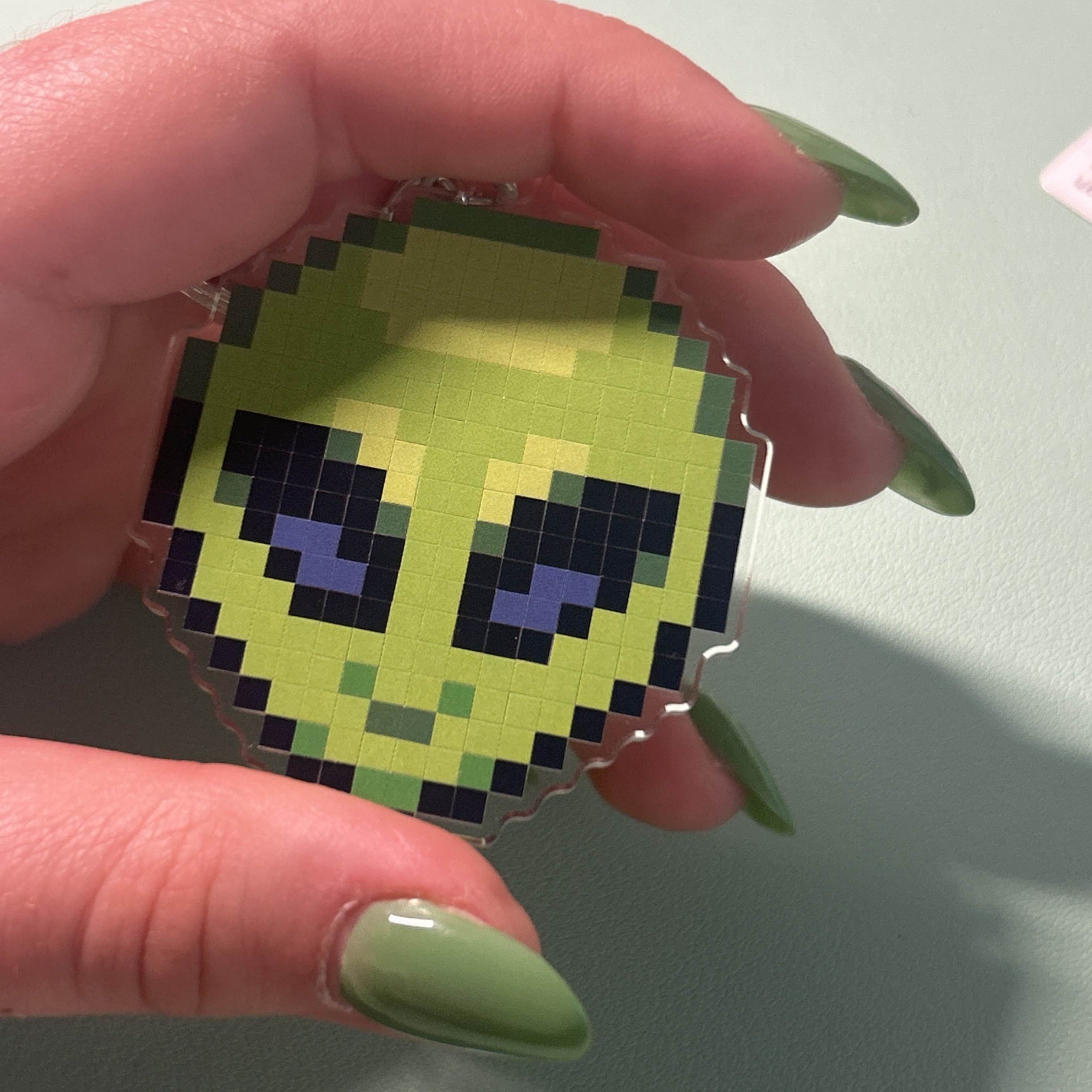 Pixelated green character held by a hand with green nail polish on a light gray background