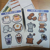 Sticker sheet with pixel art designs of coffee-themed items from Blue Hole Cafe on a wooden surface.